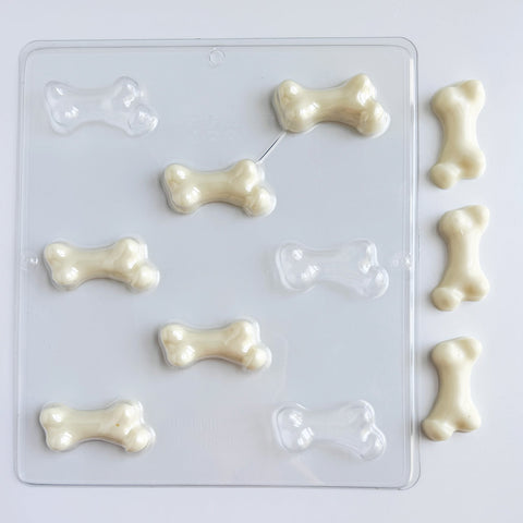 A clear plastic bone-shaped candy mold with several white chocolate bone pieces arranged beside it on a plain white background.