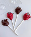 Four anatomical heart-shaped lollipops, two dark chocolate and two red with gray shading, arranged on a white surface with a clear plastic candy mold partially visible in the top left.