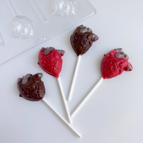 Four anatomical heart-shaped lollipops, two dark chocolate and two red with gray shading, arranged on a white surface with a clear plastic candy mold partially visible in the top left.