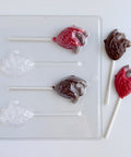A clear plastic lollipop mold shown on a white background, alongside two finished lollipops shaped like anatomical hearts in red and dark brown chocolate.