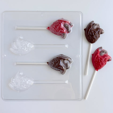 A clear plastic lollipop mold shown on a white background, alongside two finished lollipops shaped like anatomical hearts in red and dark brown chocolate.