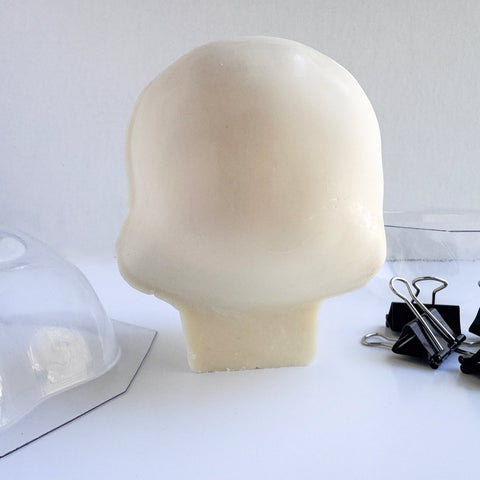 The back side of a 3d skull made from white chocolate next to the clear mold and binder clips used to make it.