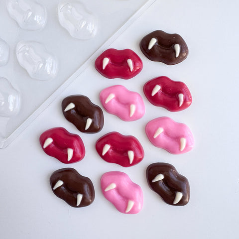 Twelve chocolate lips with fangs pieces along side the plastic candy mold used to make them. Finished chocolate pieces are shown in an assortment of red, pink, and dark chocolate with white chocolate fangs