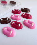 Eight vampire fang lip-shaped chocolates in pink, red, and dark chocolate, arranged on a white surface with a clear plastic chocolate mold tray in the background.