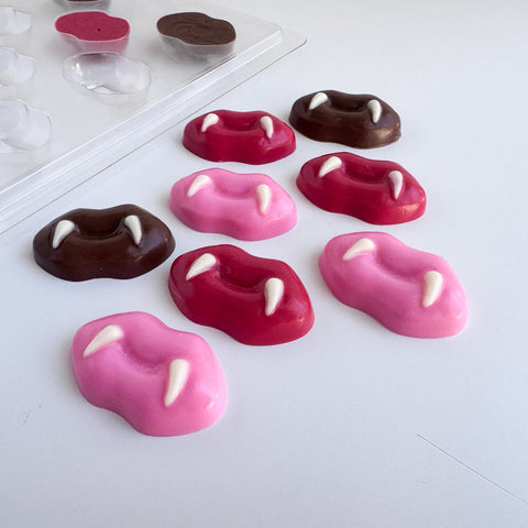 Eight vampire fang lip-shaped chocolates in pink, red, and dark chocolate, arranged on a white surface with a clear plastic chocolate mold tray in the background.
