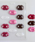 A clear plastic chocolate mold tray containing multiple lip-shaped cavities, shown alongside several finished lip-shaped chocolates with fangs in pink, red and dark chocolate. The lip designs resemble vampire or fang lips.
