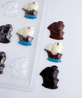 Chocolate ship candies in dark brown, milk white, and clear plastic, arranged next to a clear plastic mold tray on a white background.