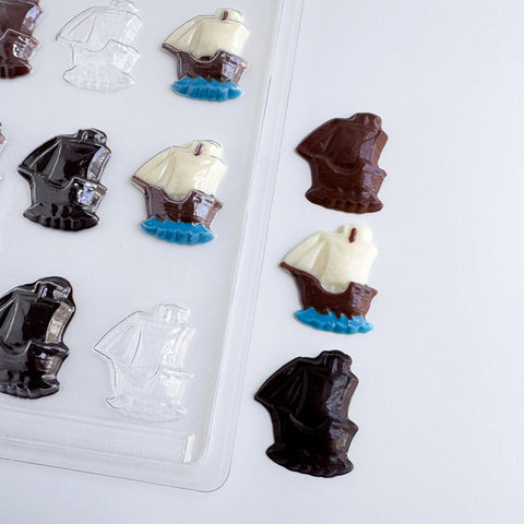 Chocolate ship candies in dark brown, milk white, and clear plastic, arranged next to a clear plastic mold tray on a white background.