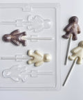 A clear plastic mummy-shaped lollipop candy mold shown on a white background, alongside two finished mummy lollipops in white chocolate and gray colored chocolate.