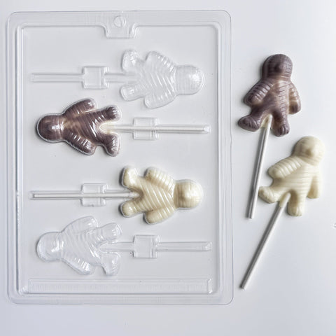 A clear plastic mummy-shaped lollipop candy mold shown on a white background, alongside two finished mummy lollipops in white chocolate and gray colored chocolate.