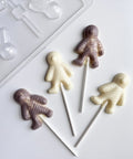 four mummy shaped lollipops in white chocolate and gray colored chocolate along side of the plastic halloween mummy chocolate mold used to make them.