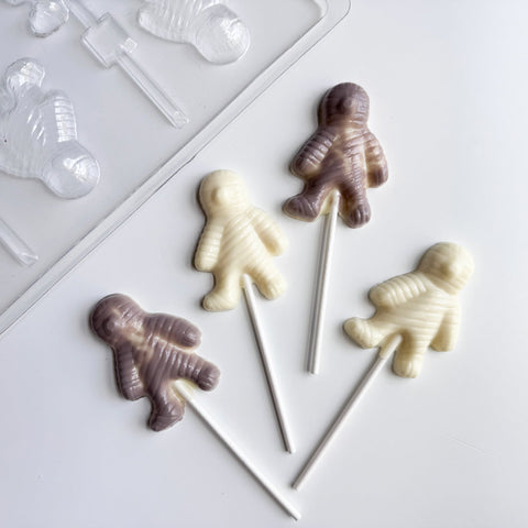 four mummy shaped lollipops in white chocolate and gray colored chocolate along side of the plastic halloween mummy chocolate mold used to make them.