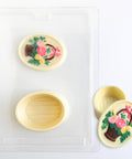 oval box made of chocolate  with a flower and basket design made using the pour box mold shown next to it