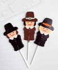 Three decorated lollipops shaped like Pilgrims on white sticks, arranged on a clean white background with the plastic mold to make them showing in the top left corner