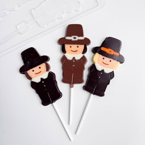 Three decorated lollipops shaped like Pilgrims on white sticks, arranged on a clean white background with the plastic mold to make them showing in the top left corner