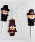 A clear plastic Thanksgiving candy mold for making pilgrim shaped lollipops. Shown along side finished chocolate lollipops. The pilgrim boy lollipops are made with milk and dark chocolate, they are wearing a hat and pilgrim clothing.