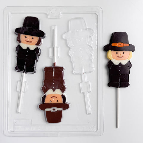 A clear plastic Thanksgiving candy mold for making pilgrim shaped lollipops. Shown along side finished chocolate lollipops. The pilgrim boy lollipops are made with milk and dark chocolate, they are wearing a hat and pilgrim clothing.