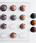 A clear plastic candy mold tray with 12 cavities, shown filled with decorated candies shaped like pumpkins. The candies feature dark chocolate brown, orange, and green details, with several finished candies displayed to the right of the mold on a clean white background.