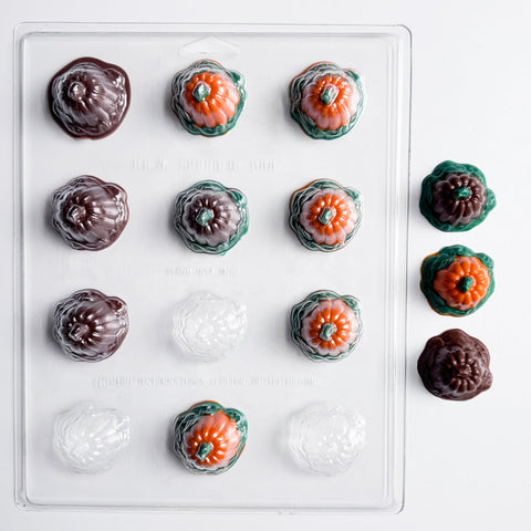 A clear plastic candy mold tray with 12 cavities, shown filled with decorated candies shaped like pumpkins. The candies feature dark chocolate brown, orange, and green details, with several finished candies displayed to the right of the mold on a clean white background.