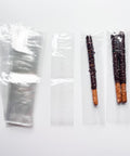 long flat cellophane bags measuring 2 1/2 inches wide by 10 inches long with a seam on one side. For packaging dipped pretzel rods or molded pretzel rods. The photo shows the clear cello bags along with one dipped pretzel in one of the cello bags and two dipped pretzels in another cello bag. The pretzels are dipped and drizzled in dark chocolate melts and sprinkled with rainbow nonpareils.