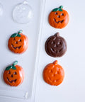 Pumpkin-shaped chocolate decorations in orange and dark brown, with green stems, arranged on a white surface next to a clear plastic candy mold.