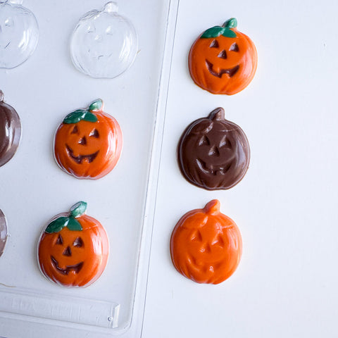 Pumpkin-shaped chocolate decorations in orange and dark brown, with green stems, arranged on a white surface next to a clear plastic candy mold.