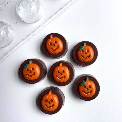 Six chocolate covered oreo cookies molded with a sandwich cookie mold with orange pumpkin faces and green leaf accents, arranged on a white surface next to the chocolate mold used to make them
