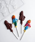 Four pumpkin scarecrow chocolate lollipops on white sticks, with two lollipops made from dark chocolate and two that are painted with colored chocolate. The finished lollipops have a pumpkin for the scarecrows head, a scarf around the neck, wearing a coat and pants, with hay coming out where the hands and feet should be, the pumpkin scarecrow is holding a lantern in it's right hand. The plastic mold used to make them is featured in the top left hand corner.
