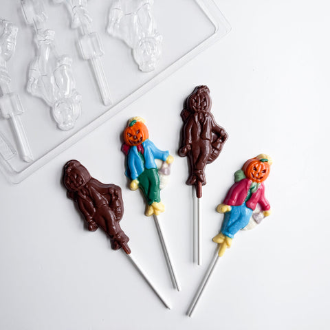 Four pumpkin scarecrow chocolate lollipops on white sticks, with two lollipops made from dark chocolate and two that are painted with colored chocolate. The finished lollipops have a pumpkin for the scarecrows head, a scarf around the neck, wearing a coat and pants, with hay coming out where the hands and feet should be, the pumpkin scarecrow is holding a lantern in it's right hand. The plastic mold used to make them is featured in the top left hand corner.