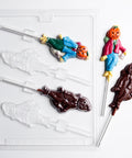 A pumpkin scarecrow lollipop mold shown along side two finished pumpkin scarecrow lollipops painted in bright colors. The plastic chocolate mold and finished chocolate lollipops are displayed on a white background.