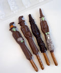 Four chocolate wizard and witch figures on pretzel sticks arranged on a white surface, with a plastic chocolate mold partially visible in the upper left corner.
