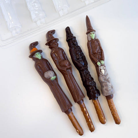 Four chocolate wizard and witch figures on pretzel sticks arranged on a white surface, with a plastic chocolate mold partially visible in the upper left corner.
