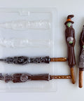 A plastic pretzel rod chocolate mold that makes spooky witch shaped lollipops with pretzel rods as the sticks. The two finished witches displayed to the right of the mold have green faces, dark chocolate cloaks and hats, one is holding a cauldron/pumpkin, and the other is holding a skeleton decoration. 
