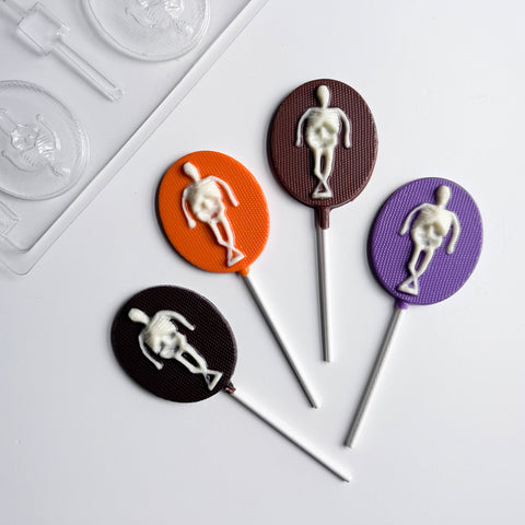 4 oval skeleton lollipop chocolates next to the plastic candy mold used to make them on a white background