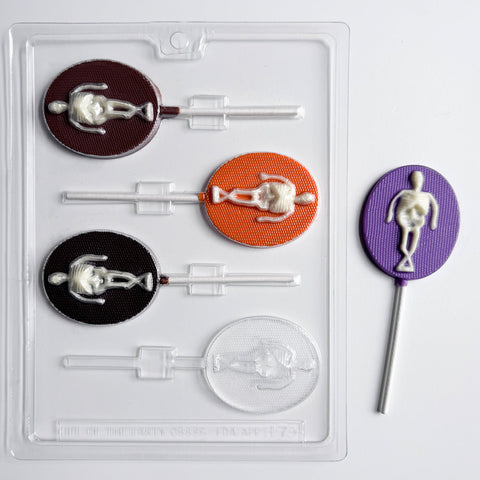 oval lollipop chocolate mold with skeleton in the center. One finished skeleton lollipop to the right of the plastic mold, made with purple chocolate for the oval part and the skeleton is made in white chocolate.