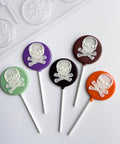 Five round lollipops with white skull and crossbones designs on colorful round chocolate backgrounds, arranged on a white surface with the plastic chocolate mold used to make them partially visible in the top left.