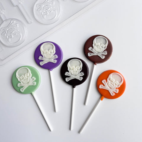 Five round lollipops with white skull and crossbones designs on colorful round chocolate backgrounds, arranged on a white surface with the plastic chocolate mold used to make them partially visible in the top left.