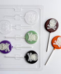 skull and crossbones lollipop chocolate mold that makes round lollipops with skull and crossbones imprinted in the center. Two finished lollipops to the right of the mold are colored in orange and milk chocolate with white skulls, placed next to the plastic candy mold.