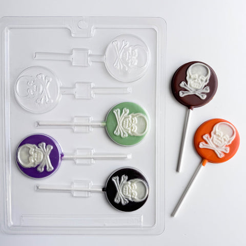skull and crossbones lollipop chocolate mold that makes round lollipops with skull and crossbones imprinted in the center. Two finished lollipops to the right of the mold are colored in orange and milk chocolate with white skulls, placed next to the plastic candy mold.