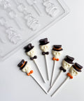 Six white chocolate skull pops with dark chocolate top hats, black bow ties, and orange bow ties on white lollipop sticks, arranged on a white surface with a clear plastic skull with hat lollipop candy mold in the background.