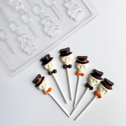 Six white chocolate skull pops with dark chocolate top hats, black bow ties, and orange bow ties on white lollipop sticks, arranged on a white surface with a clear plastic skull with hat lollipop candy mold in the background.