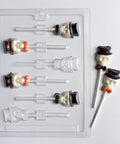 A clear plastic Halloween-themed skull with hat lollipop mold shown with two finished lollipops on a white background. The lollipops feature a white chocolate skull wearing a black top hat and orange bow tie.