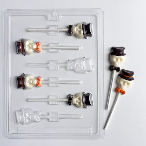 A clear plastic Halloween-themed skull with hat lollipop mold shown with two finished lollipops on a white background. The lollipops feature a white chocolate skull wearing a black top hat and orange bow tie.