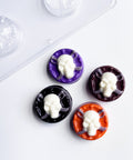 four oreo cookies molded with chocolate sculls using the skull with swords sandwich cookie mold. The finished molded sandwich cookies have dark, black, purple, and orange chocolate bases with white skulls on top and gray swords behind the skulls. All displayed along side the plastic chocolate mold used to make them.
