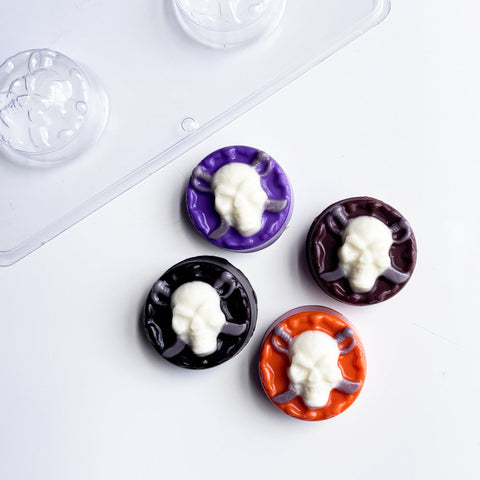 four oreo cookies molded with chocolate sculls using the skull with swords sandwich cookie mold. The finished molded sandwich cookies have dark, black, purple, and orange chocolate bases with white skulls on top and gray swords behind the skulls. All displayed along side the plastic chocolate mold used to make them.