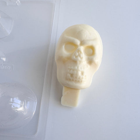 Plastic skull candy mold next to a finished white chocolate 3d skull that has a chocolate base at the bottom to insert into a cake and use as a topper, all sitting on a white back drop.