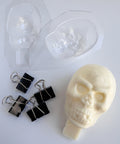 A 3D skull made from white chocolate sitting next to the clear plastic candy mold and binder clips used to mold it. The white chocolate skull has a base at the bottom that can be inserted into a cake to act as a cake topper.
