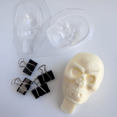 A 3D skull made from white chocolate sitting next to the clear plastic candy mold and binder clips used to mold it. The white chocolate skull has a base at the bottom that can be inserted into a cake to act as a cake topper.