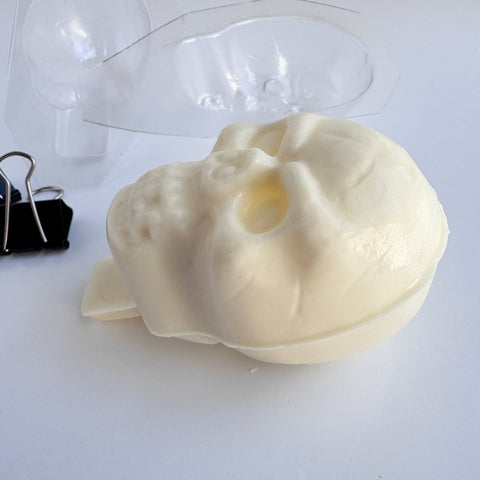 Plastic skull candy mold next to a finished white chocolate 3d skull that has a chocolate base at the bottom to insert into a cake and use as a topper.
Binder clips that were used for clamping the mold together are shown on the left.