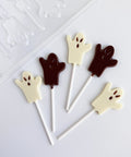 Five ghost-shaped lollipops in white and dark chocolate, arranged on a white surface with a clear plastic candy mold partially visible in the top left corner.